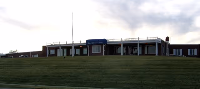 Wide view of a single-story brick building with a sign that reads 'Blue Hill Care Center' on the front. The building is situated on a grassy hill under a cloudy sky.