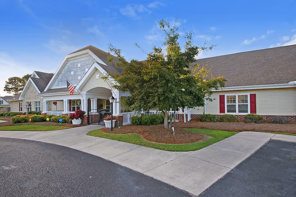 Exterior view of The Pines at Florence Assisted Living & Memory Care building with a well-maintained garden, a tree, and an American flag near the entrance under a clear blue sky.