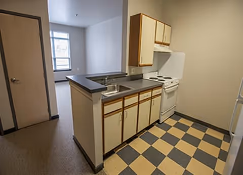 Small kitchen area with checkered black and beige floor tiles, white cabinets with wooden trim, a white stove, and a sink with a countertop. Adjacent room with a window and beige carpet is visible through an open doorway.