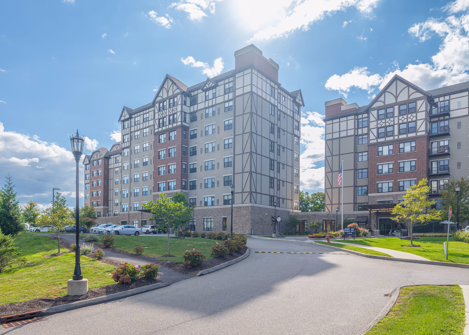 Exterior view of a multi-story senior living facility building with Tudor-style architectural elements, surrounded by landscaped greenery, a driveway, and a clear blue sky with some clouds.