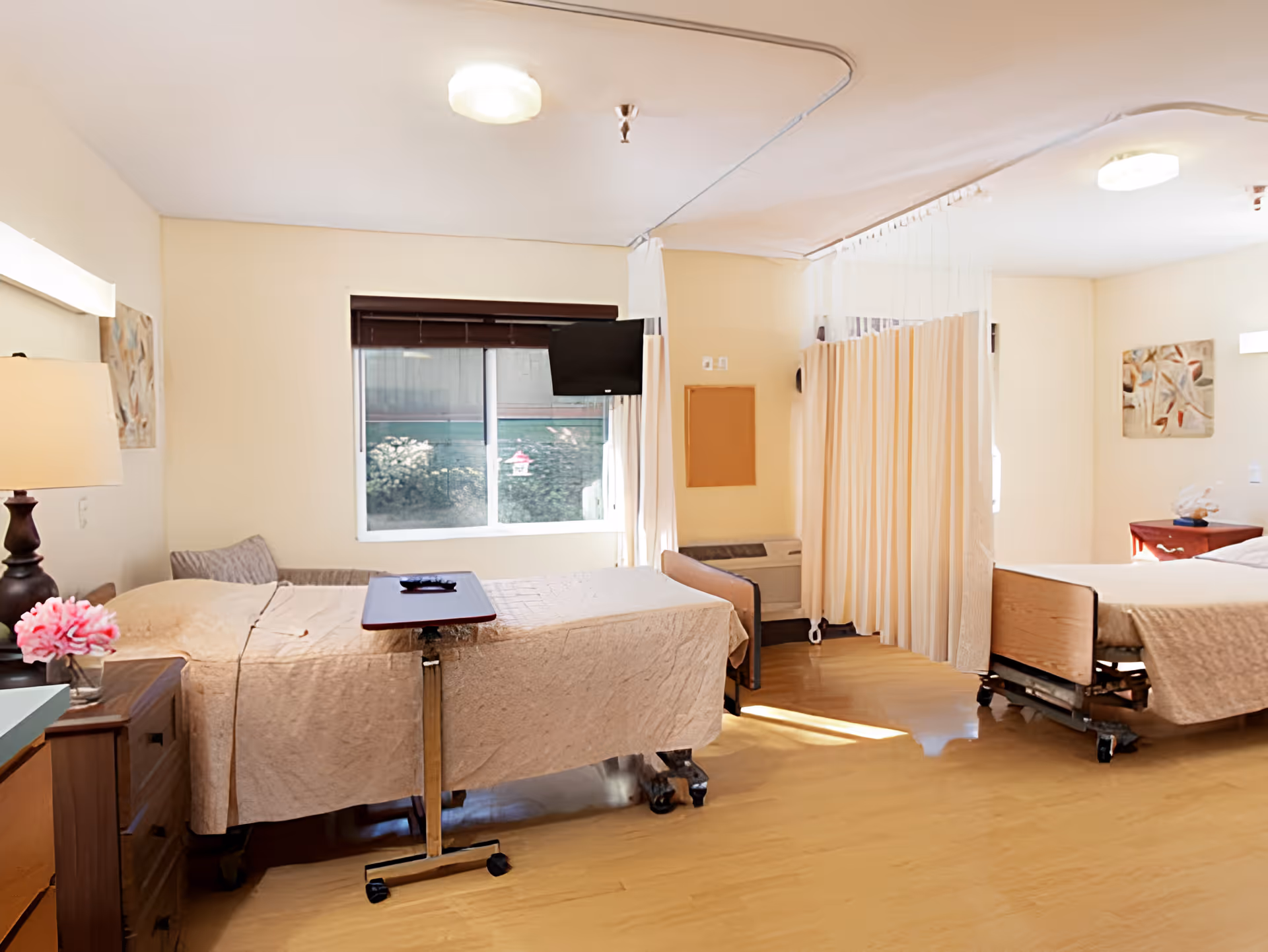 A bright room with two hospital-style beds separated by a beige privacy curtain. Each bed has a beige quilted blanket and a small overbed table. There is a window with a mounted TV on the wall between the beds. The room has light-colored walls, wooden flooring, and bedside tables with lamps and decorative items.