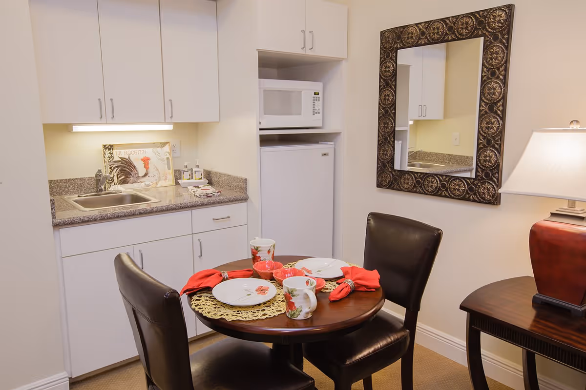 A small kitchenette area with white cabinets, a countertop with a sink, a microwave, and a mini refrigerator. In front of the kitchenette is a round wooden table set with two plates, mugs, and red napkins, accompanied by two dark leather chairs. A decorative mirror hangs on the wall above a side table with a red lamp.