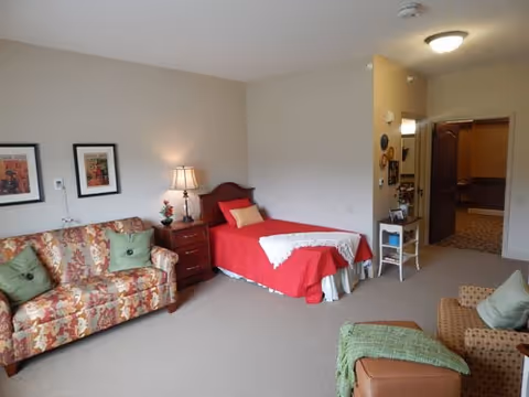 A cozy senior living room with a single bed covered in a red bedspread and a white lace throw, a floral patterned sofa with green pillows, a wooden nightstand with a lamp and flower vase, a wicker chair with a green throw, and a small ottoman. The room has beige walls and carpet, with framed artwork on the wall and an open doorway leading to another room.