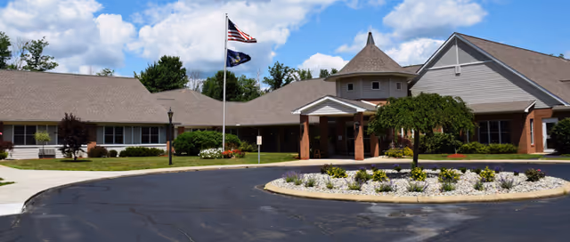 Exterior view of a single-story senior living facility building with a circular driveway and landscaped roundabout featuring small plants and rocks. Two flagpoles with the American flag and another flag are visible near the entrance. The sky is partly cloudy.