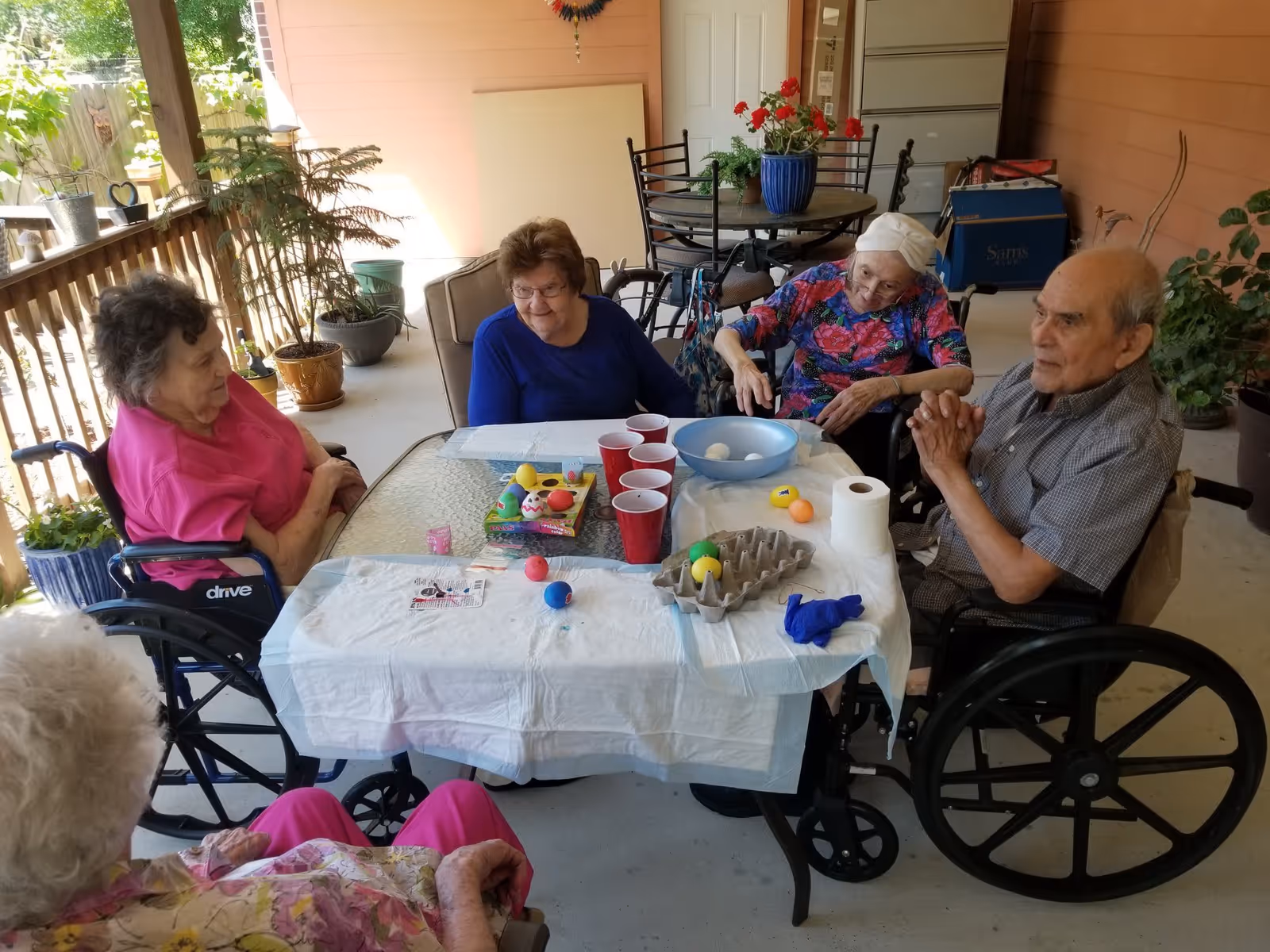 A group of elderly people sitting around a table on a covered outdoor patio. They appear to be engaged in an activity involving colorful plastic eggs and cups. The area is decorated with potted plants and outdoor furniture.