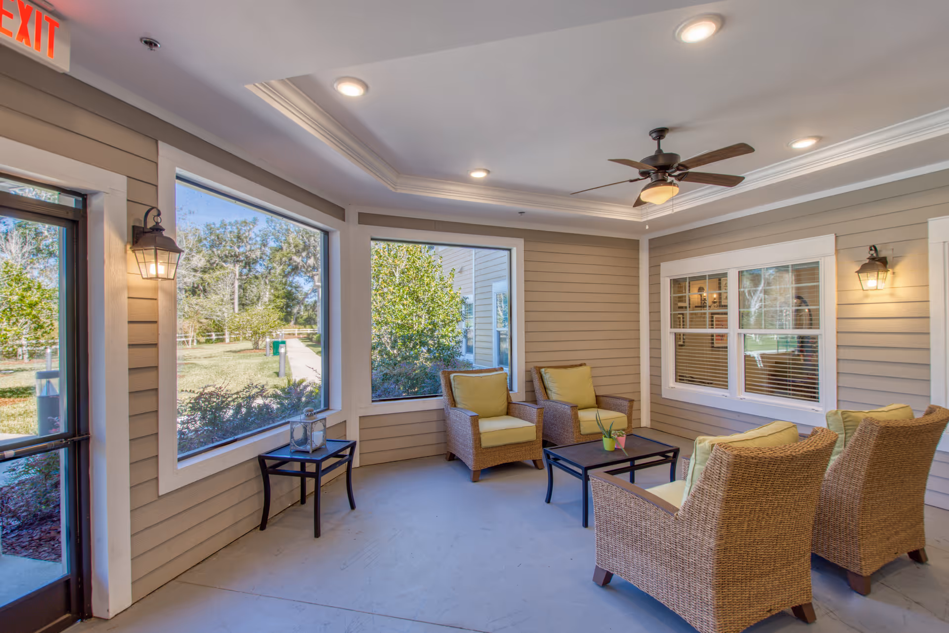 A cozy indoor seating area with four wicker armchairs with yellow cushions arranged around a black coffee table. The room has large windows showing a view of greenery outside, beige walls with white trim, ceiling lights, a ceiling fan, and two wall-mounted lantern-style lights. An exit door is visible on the left side.