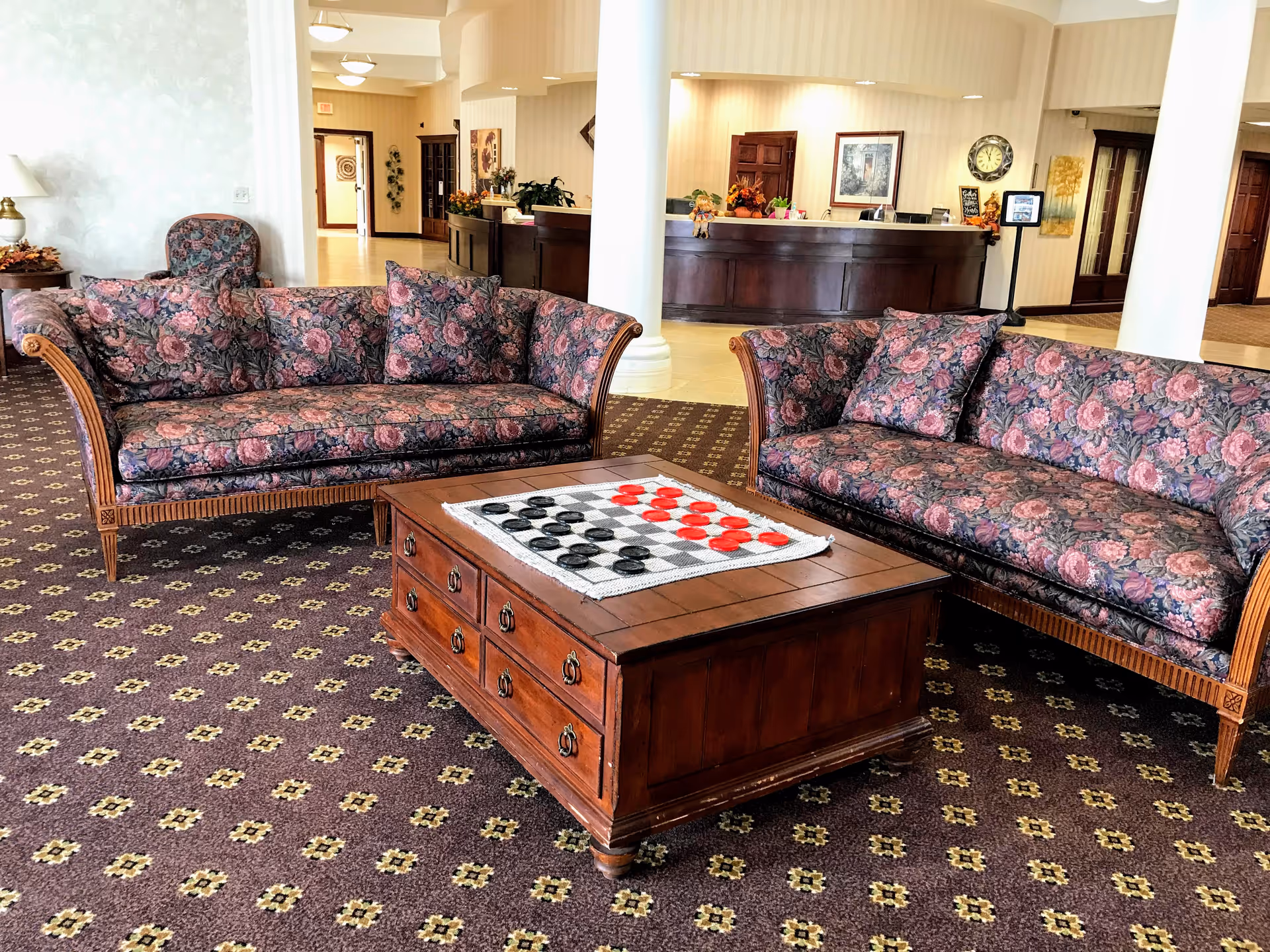 A senior living lobby with two floral sofas facing a wooden coffee table topped with a checkerboard and a reception desk in the background.