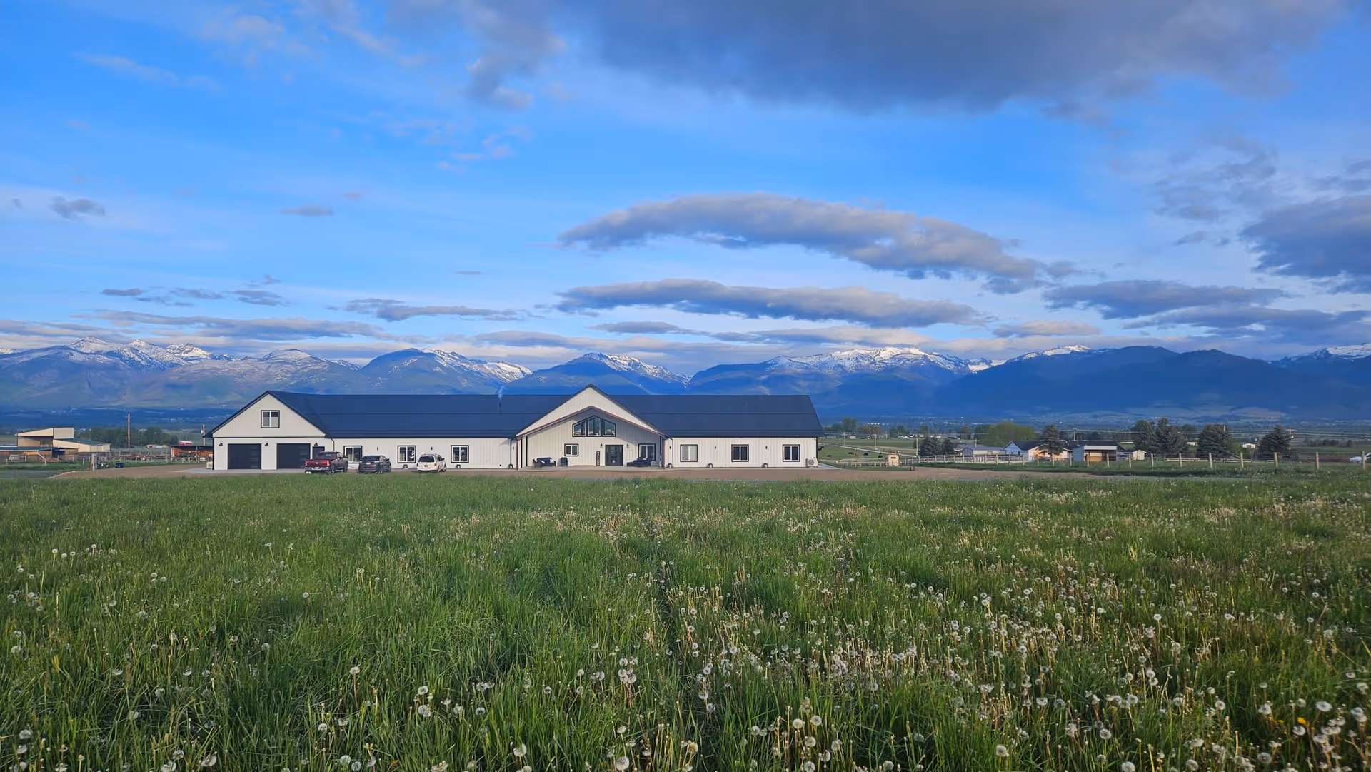 A wide view of a single-story building with a dark roof and white walls, situated in a large grassy field with dandelions. In the background, there are snow-capped mountains under a partly cloudy blue sky.