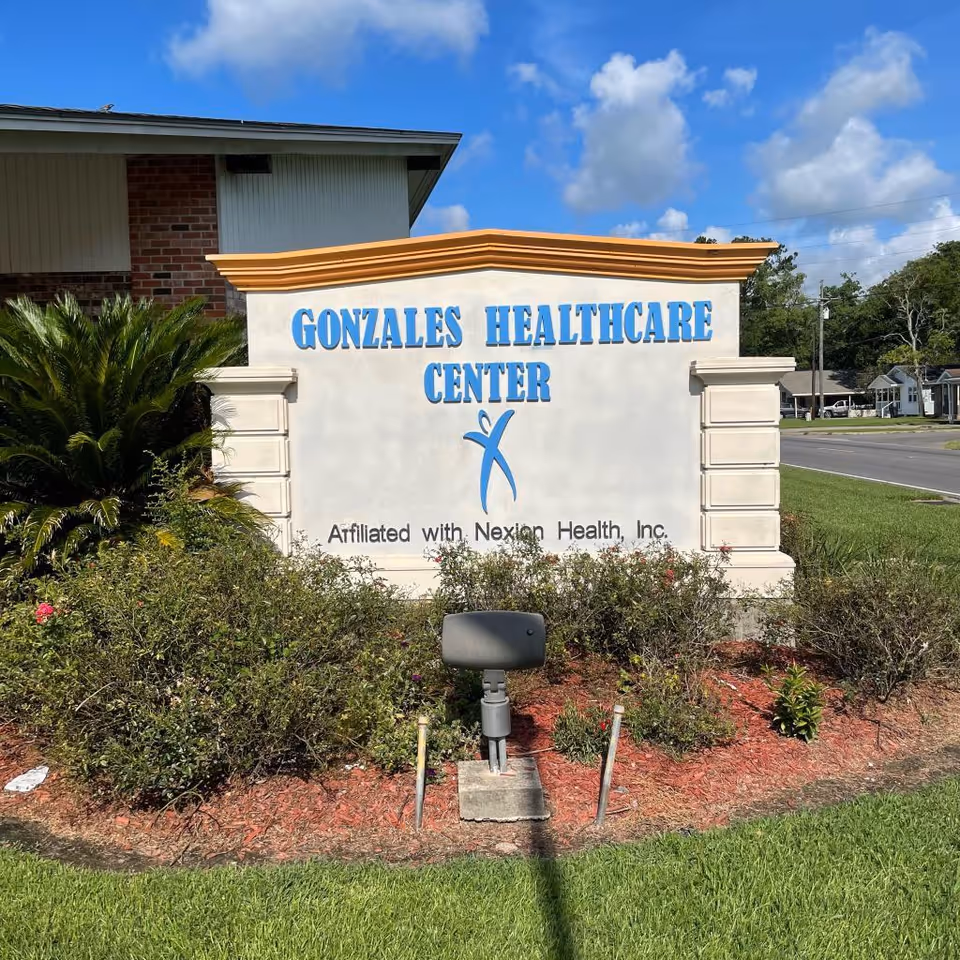 Outdoor sign for Gonzales Healthcare Center with blue text and a logo, surrounded by landscaping with bushes and grass, under a partly cloudy blue sky.