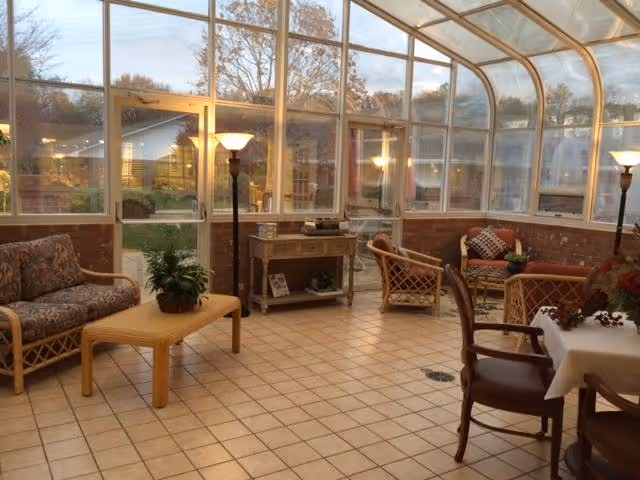 A bright sunroom with large windows and a glass ceiling, furnished with wicker chairs, a wicker sofa with floral cushions, a wooden coffee table with a potted plant, a small wooden console table, floor lamps, and a dining table with chairs. Outside, trees and houses are visible through the windows.