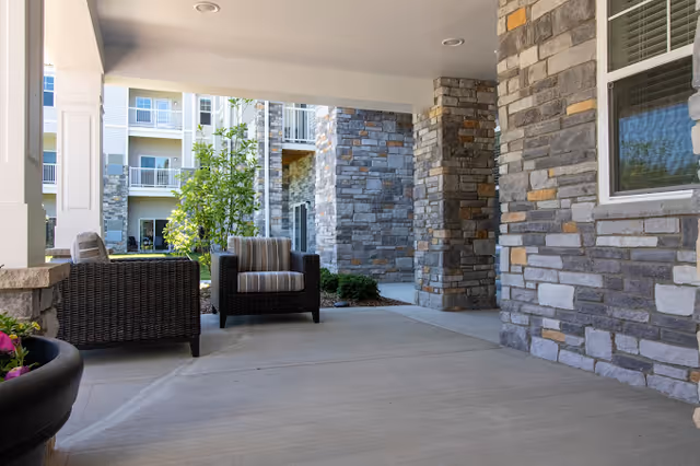 Covered outdoor patio area with stone pillars and two wicker chairs with cushions. The background shows part of a multi-story building with balconies and greenery.