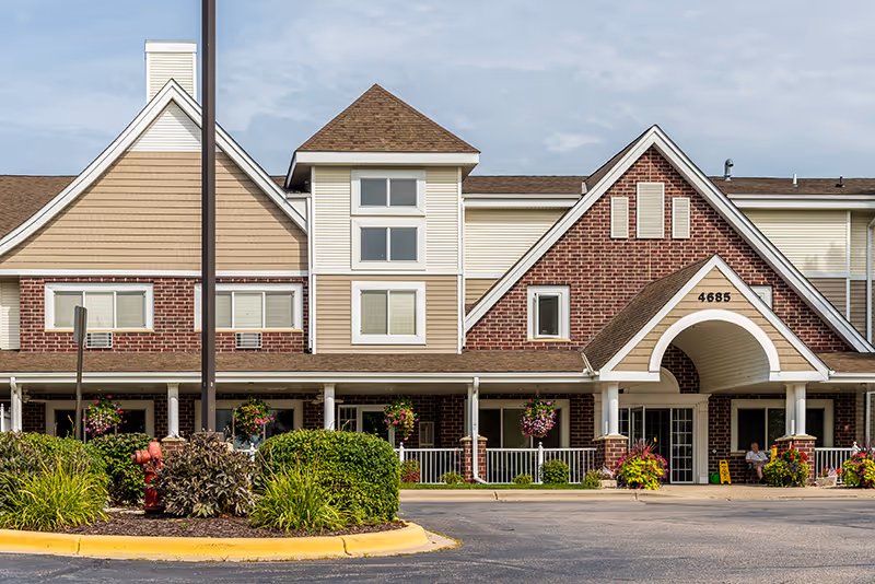 Front exterior view of a senior living facility building with brick and beige siding, a peaked roof, and a covered entrance with the number 4685 above it. There are hanging flower baskets and landscaped bushes in front of the building, with a person sitting near the entrance.