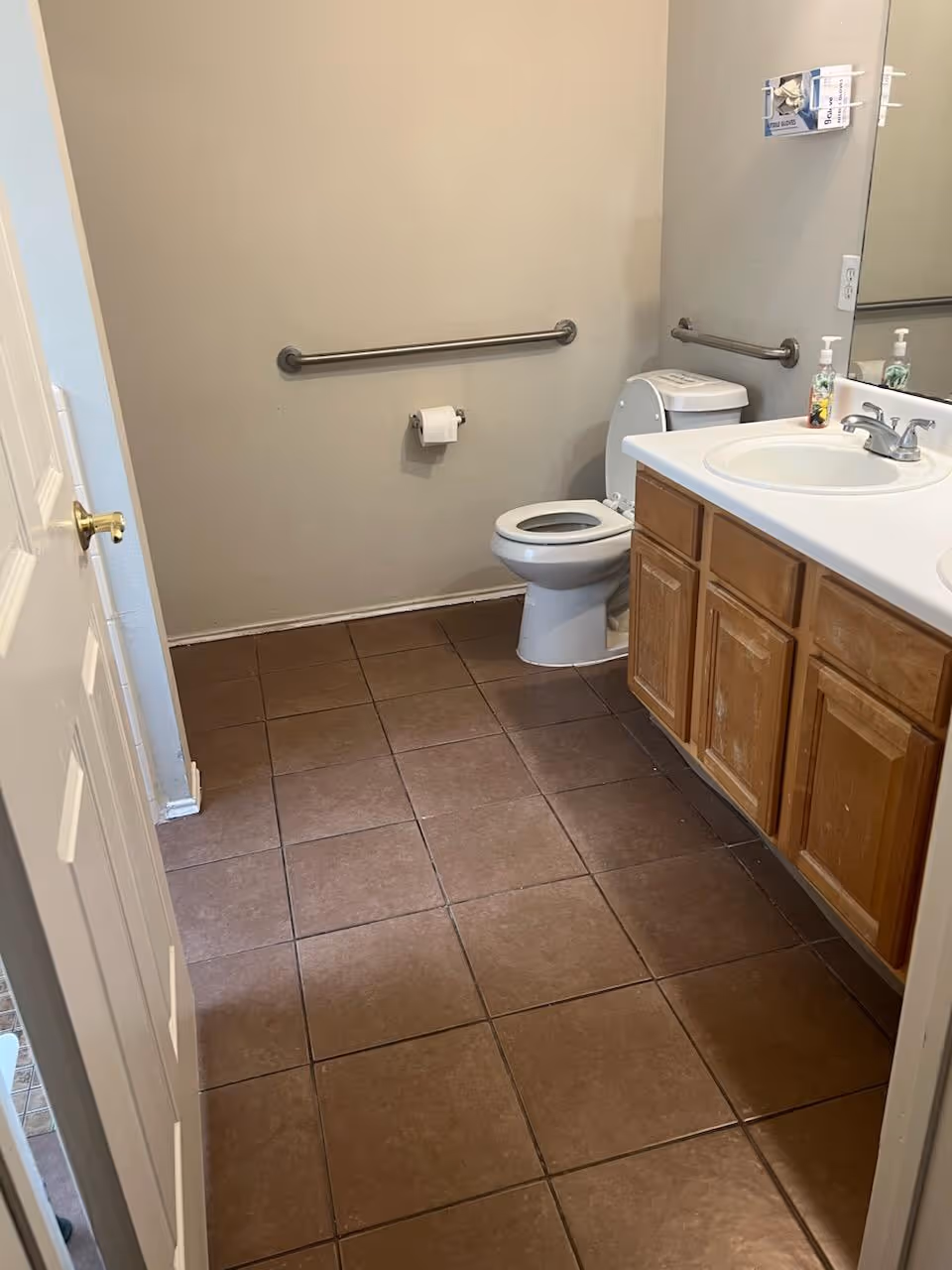 A bathroom with brown tiled floor, a white toilet with the lid open, a white sink with a silver faucet on a wooden cabinet, a large mirror above the sink, a soap dispenser, and a grab bar on the wall next to the toilet and under the mirror.