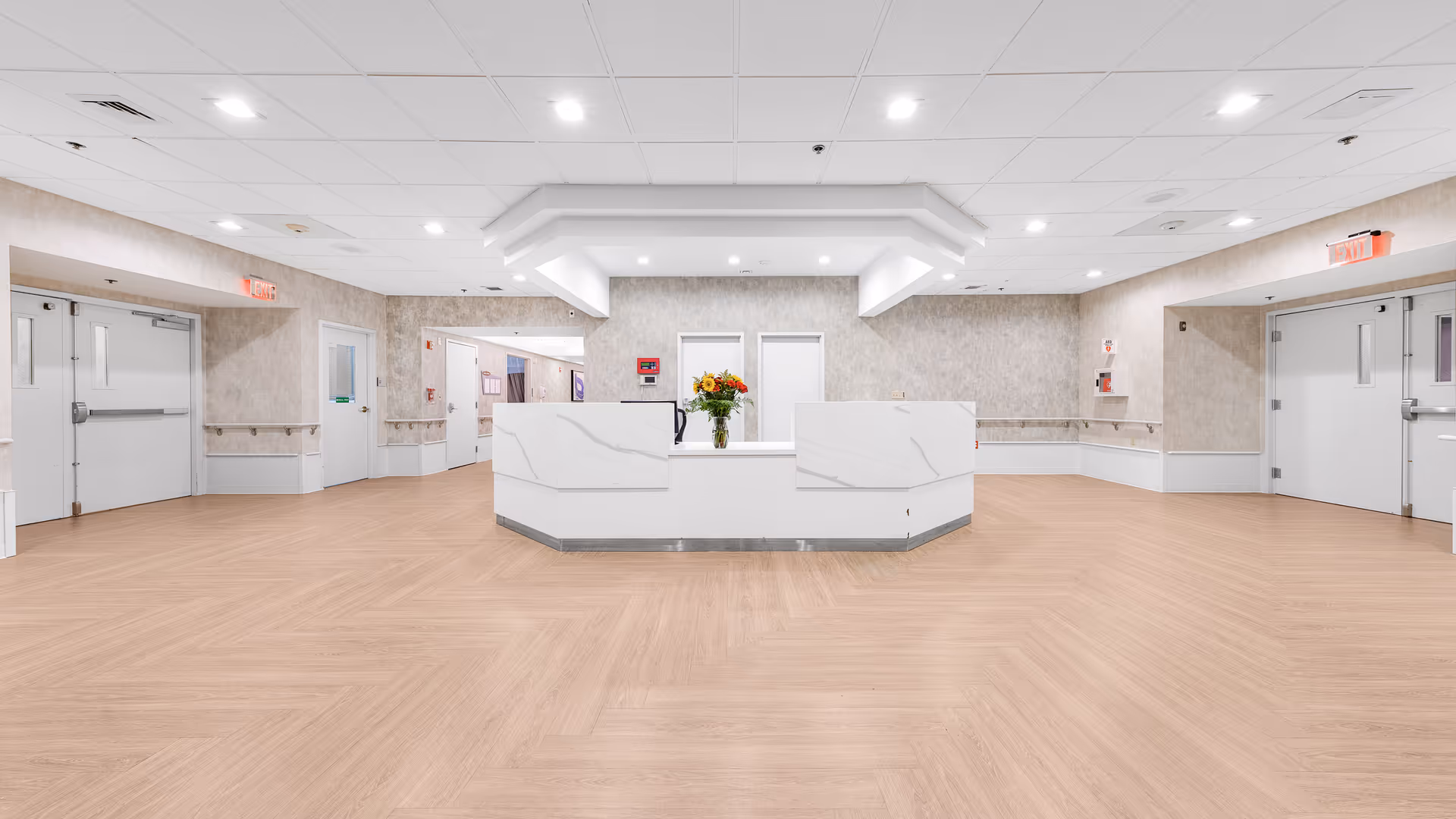 Bright, spacious reception desk with a vase of flowers in a modern facility hallway with light wood floors and overhead lighting.