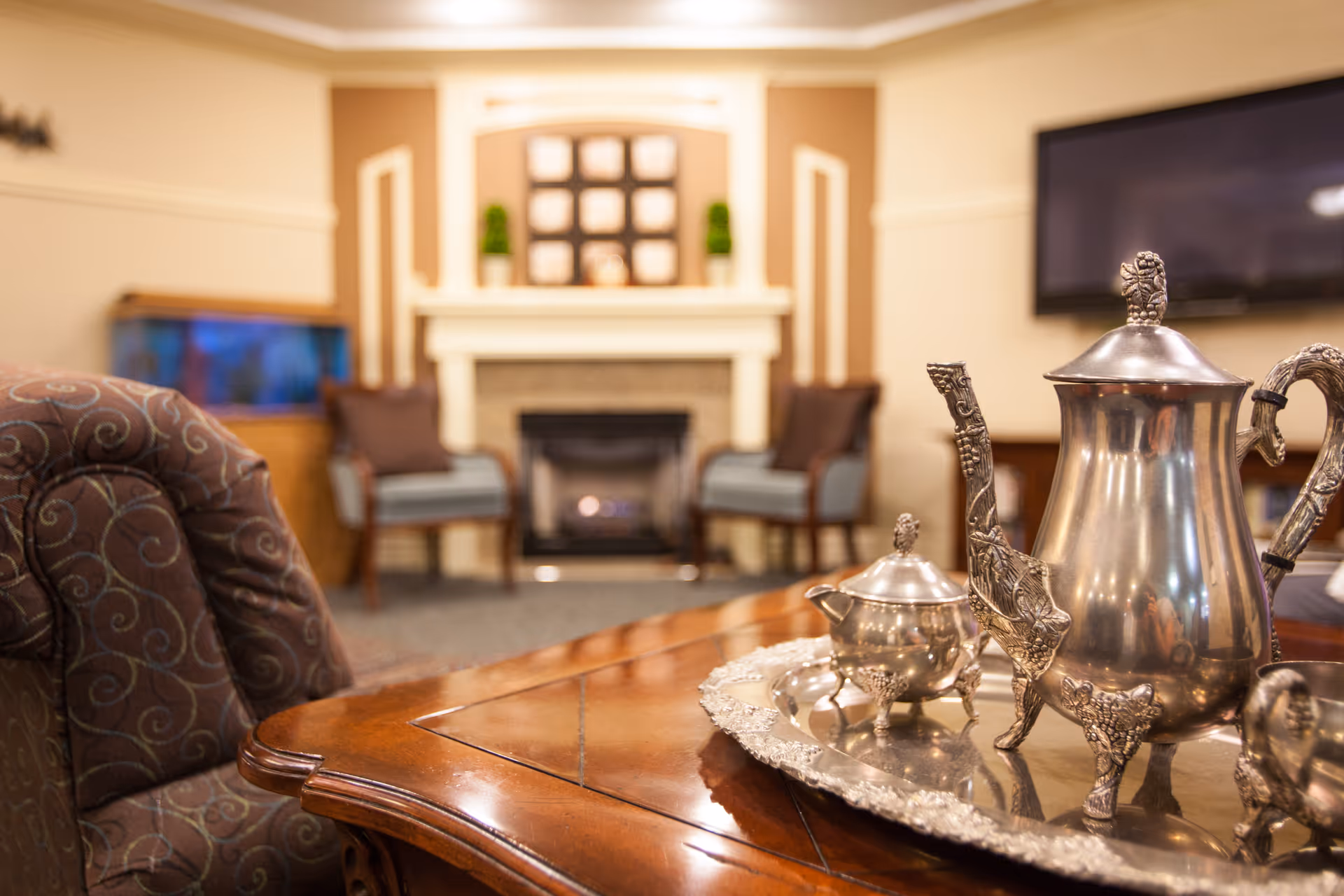 A cozy living room area with a wooden coffee table in the foreground holding an ornate silver tea set. In the background, there are two armchairs with cushions, a fireplace with decorative items on the mantel, a mounted flat-screen TV, and a fish tank to the left.