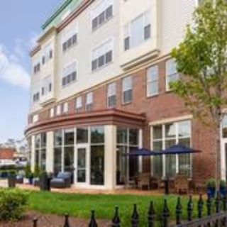 Exterior front of a multi-story senior living building with large windows, a patio with umbrellas and seating facing a small lawn.