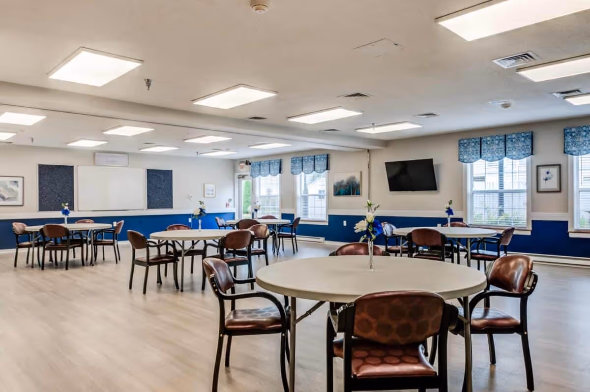Bright communal dining/activity room with round tables and chairs, a wall-mounted TV, and windows with blue valances.