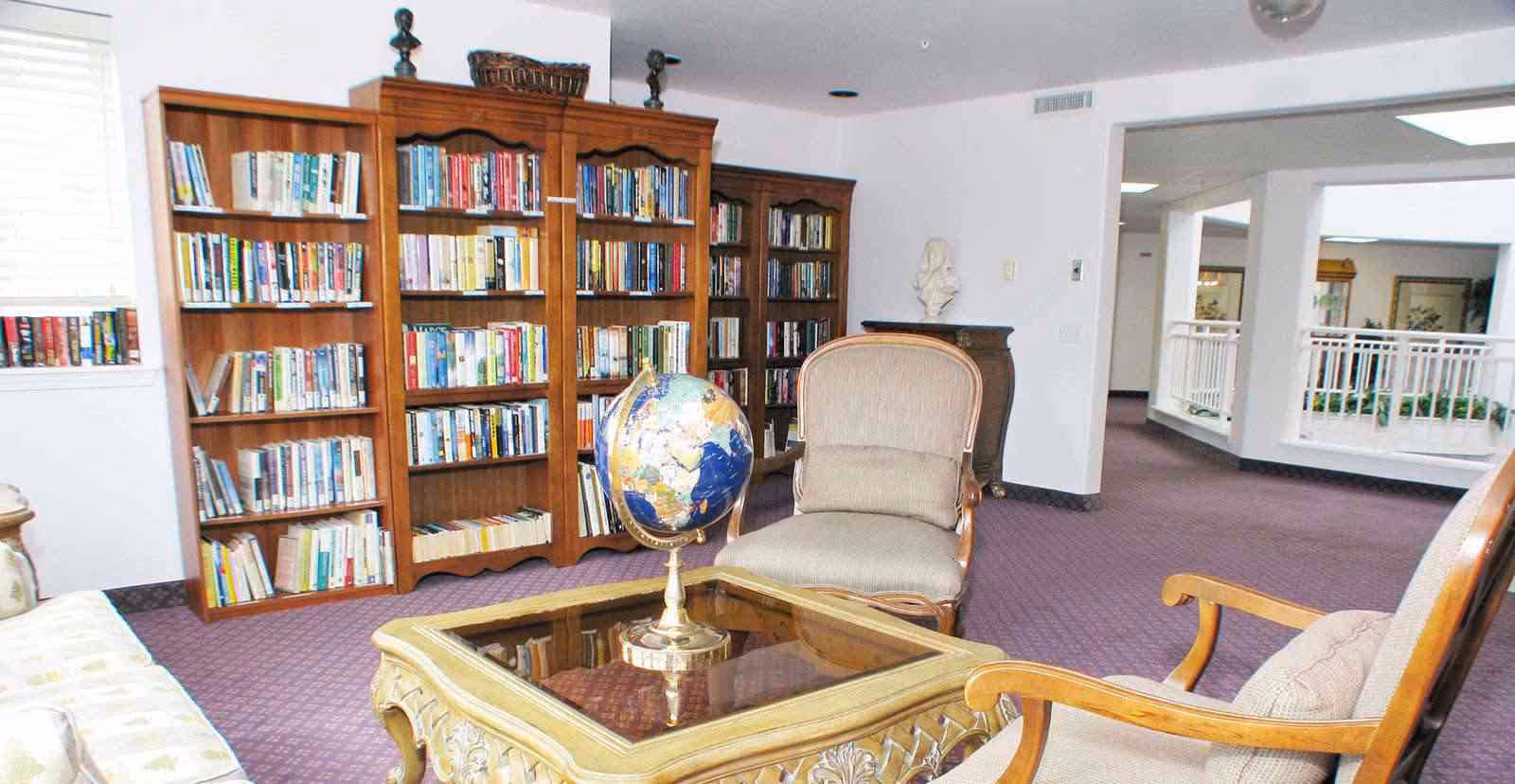 A cozy reading area in a senior living facility featuring wooden bookshelves filled with books, two upholstered armchairs, a glass-top wooden coffee table with a globe on it, and a purple carpet. The space opens into a hallway with white railings and natural light.