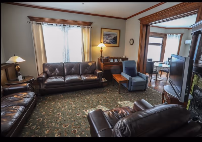 A cozy living room with dark brown leather sofas and armchairs arranged around a green patterned carpet. There is a wooden side table with a lamp on the left, a small wooden table with a blue armchair in the center, and a flat-screen TV on a stand near a fireplace on the right. The room has cream-colored walls with wooden trim, a window with light curtains, and an adjacent dining area visible through a large wooden-framed opening.