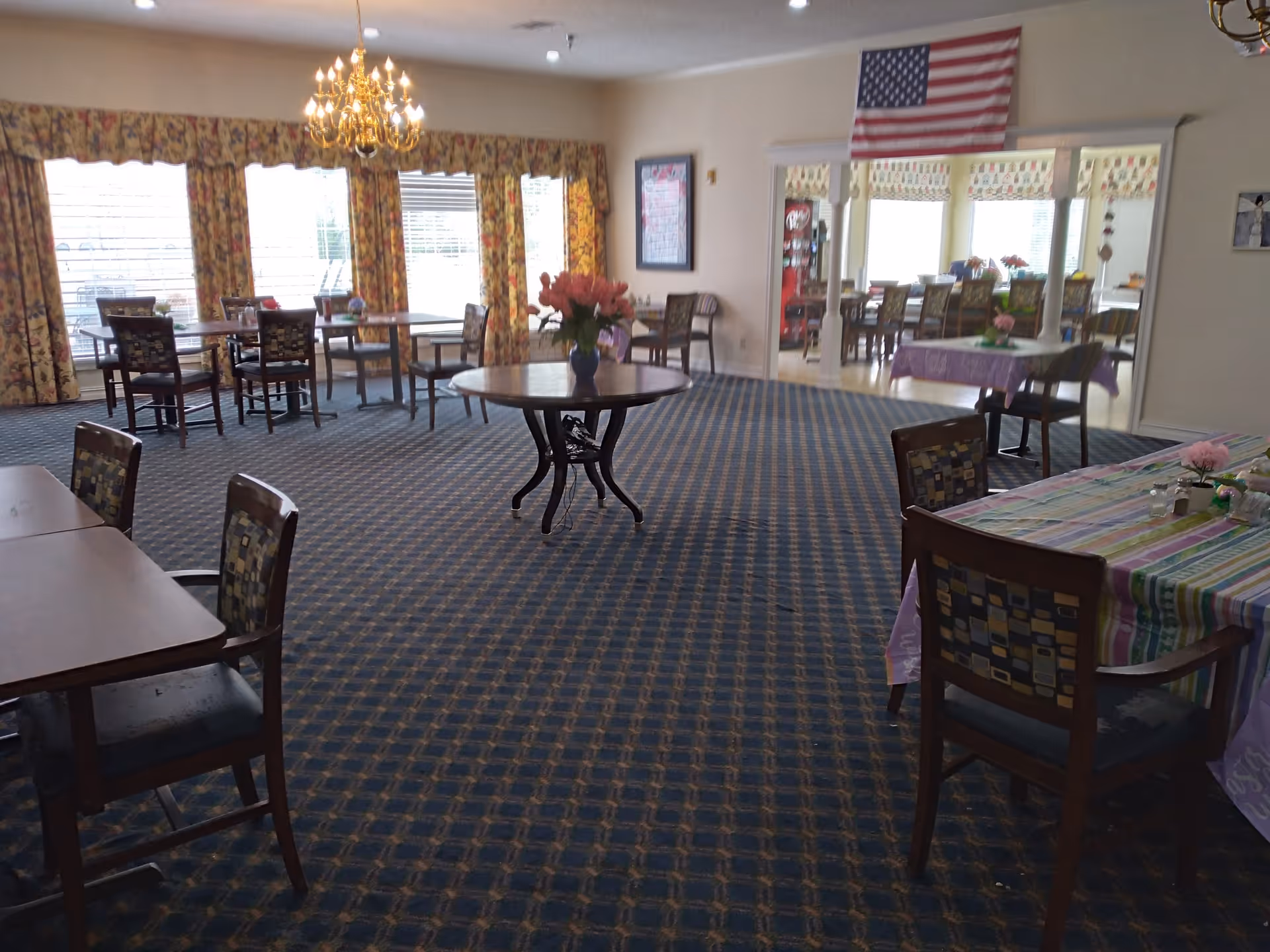 A dining room in an assisted living community with multiple tables and chairs arranged on a patterned carpet. The room has large windows with floral curtains, a chandelier hanging from the ceiling, and an American flag displayed on the wall. There are flowers in vases on some tables and a glimpse of another room with more tables and chairs through an open doorway.