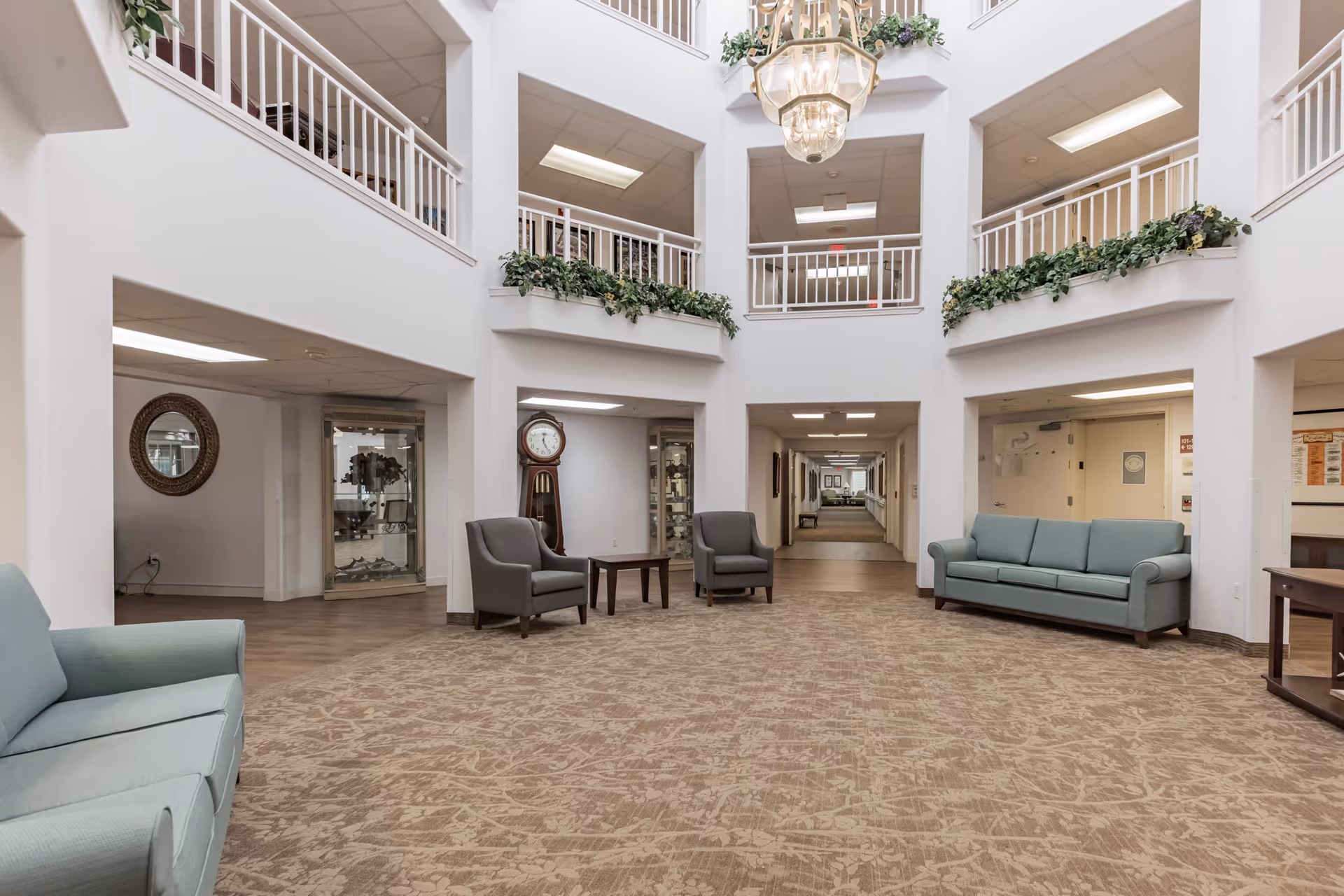 Spacious and well-lit common area in a senior living facility with beige patterned carpet, two light blue sofas, two gray armchairs, a wooden coffee table, a grandfather clock, and decorative plants on the upper balcony railings. The area has white walls and a large hanging chandelier, with a hallway leading further into the building.