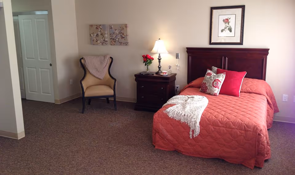 A cozy bedroom with a single bed covered in a coral quilt and decorative pillows. Next to the bed is a dark wooden nightstand with a lamp, a vase of red roses, and some small items. A beige armchair with a pink throw is placed against the wall, which is decorated with two floral paintings. The room has beige walls and carpeted flooring.