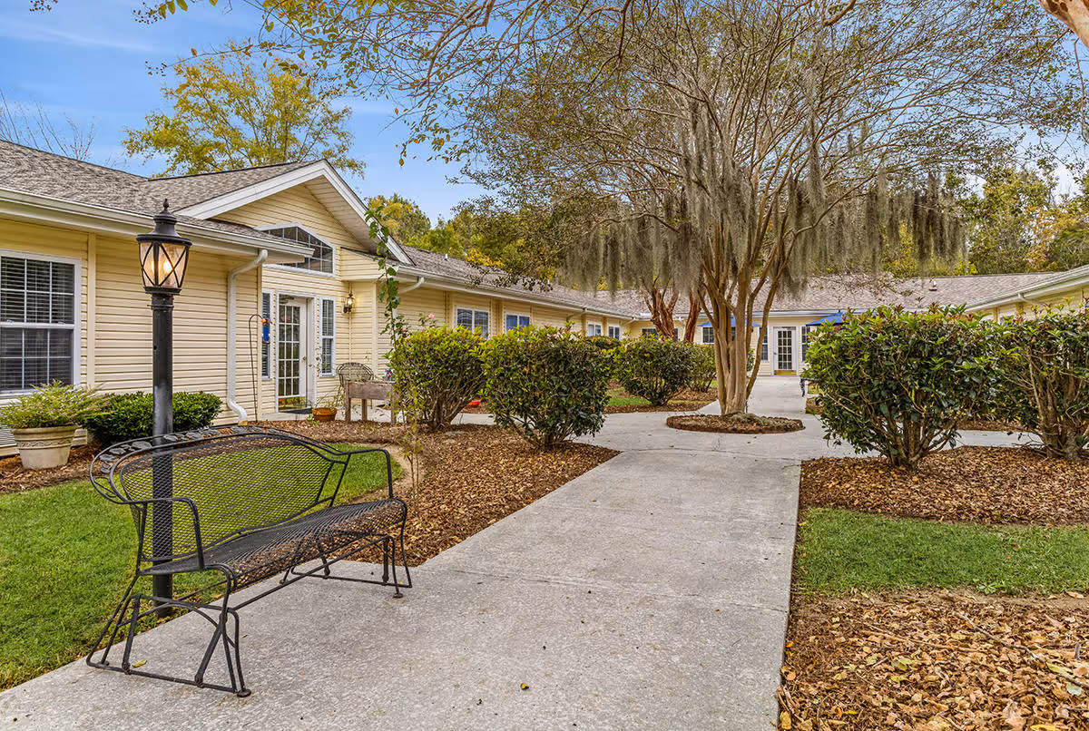 Outdoor courtyard area at Pinewood Square featuring a paved walkway, a black metal bench, a lamp post, trimmed bushes, and trees with hanging moss, surrounded by a single-story building with beige siding and white-framed windows and doors.