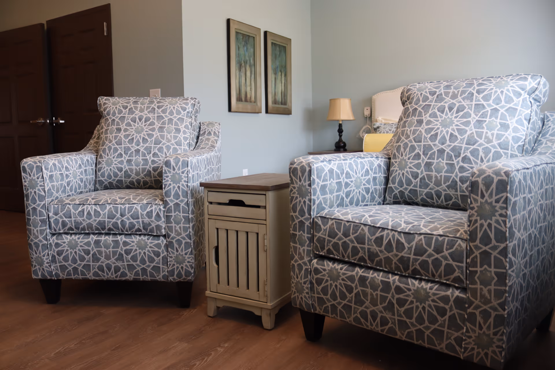 Two patterned armchairs flanking a small wooden side table in a softly lit living area with a lamp and wall art.