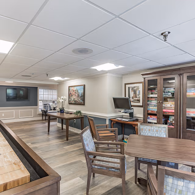 A well-lit common area in a senior living facility featuring wooden tables and chairs with patterned cushions. There is a cabinet filled with board games, a computer workstation, and framed artwork on the walls. The room has a light-colored ceiling with recessed lighting and wood-style flooring.