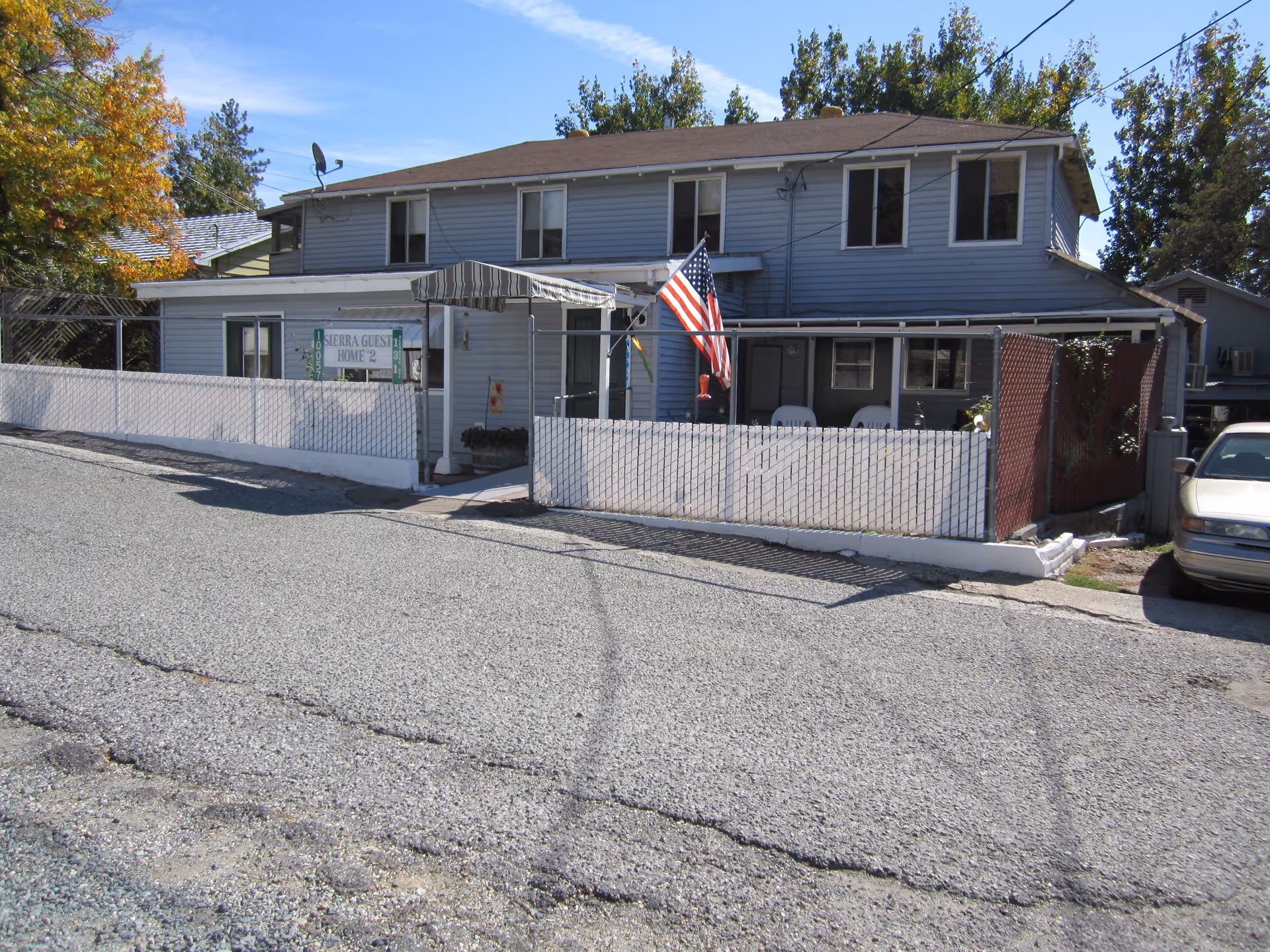 Exterior view of a two-story residential building with light blue siding, a brown roof, and a fenced front porch area. An American flag is displayed near the entrance, and there is a sign that reads 'Sierra Guest Home #2'. A car is parked on the right side of the building, and trees with autumn foliage are visible in the background.