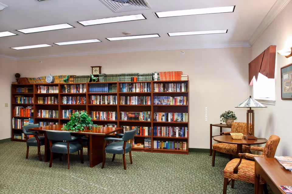 Well-lit library-style common room with bookshelves, a central table surrounded by chairs, and a small seating area with armchairs and a lamp.