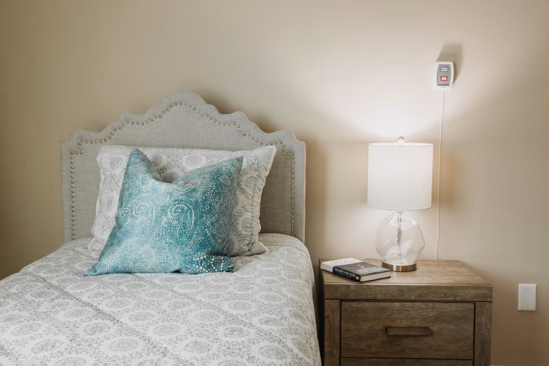 A neatly made bed with a decorative headboard and two pillows, one white and one teal with a paisley pattern. Next to the bed is a wooden nightstand with a clear glass lamp and a book titled 'Reason for Hope'. The wall behind is beige with a mounted emergency call button above the nightstand.