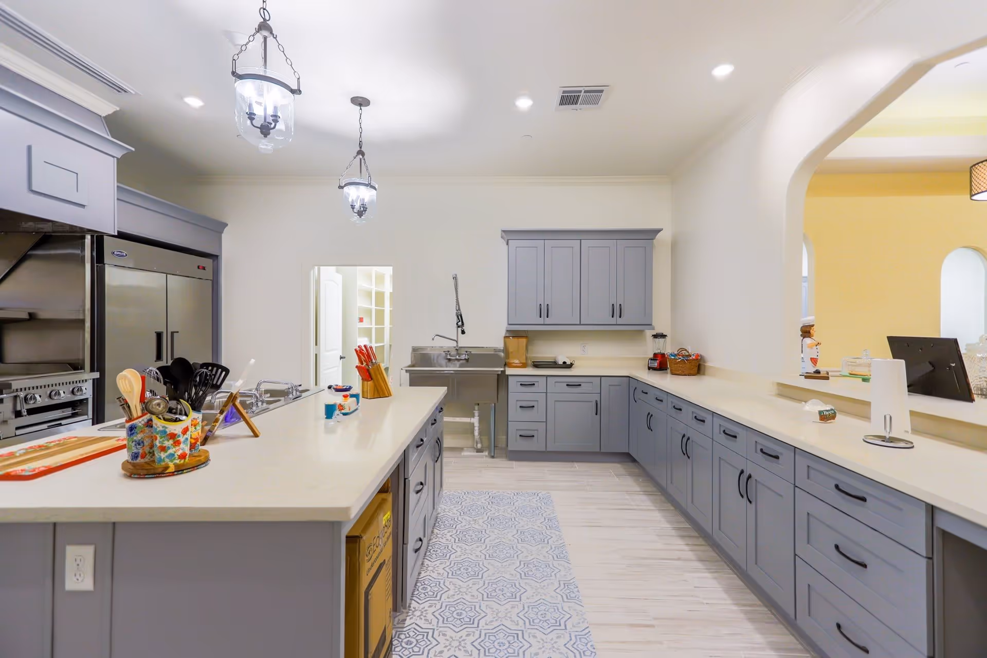 A spacious, modern kitchen with light gray cabinets and white countertops. The kitchen features a large island with various kitchen utensils, a knife block, and a colorful container. Stainless steel appliances including a refrigerator and stove are visible on the left. Two pendant lights hang from the ceiling, and there is a patterned rug on the floor. An open archway leads to another room with yellow walls.