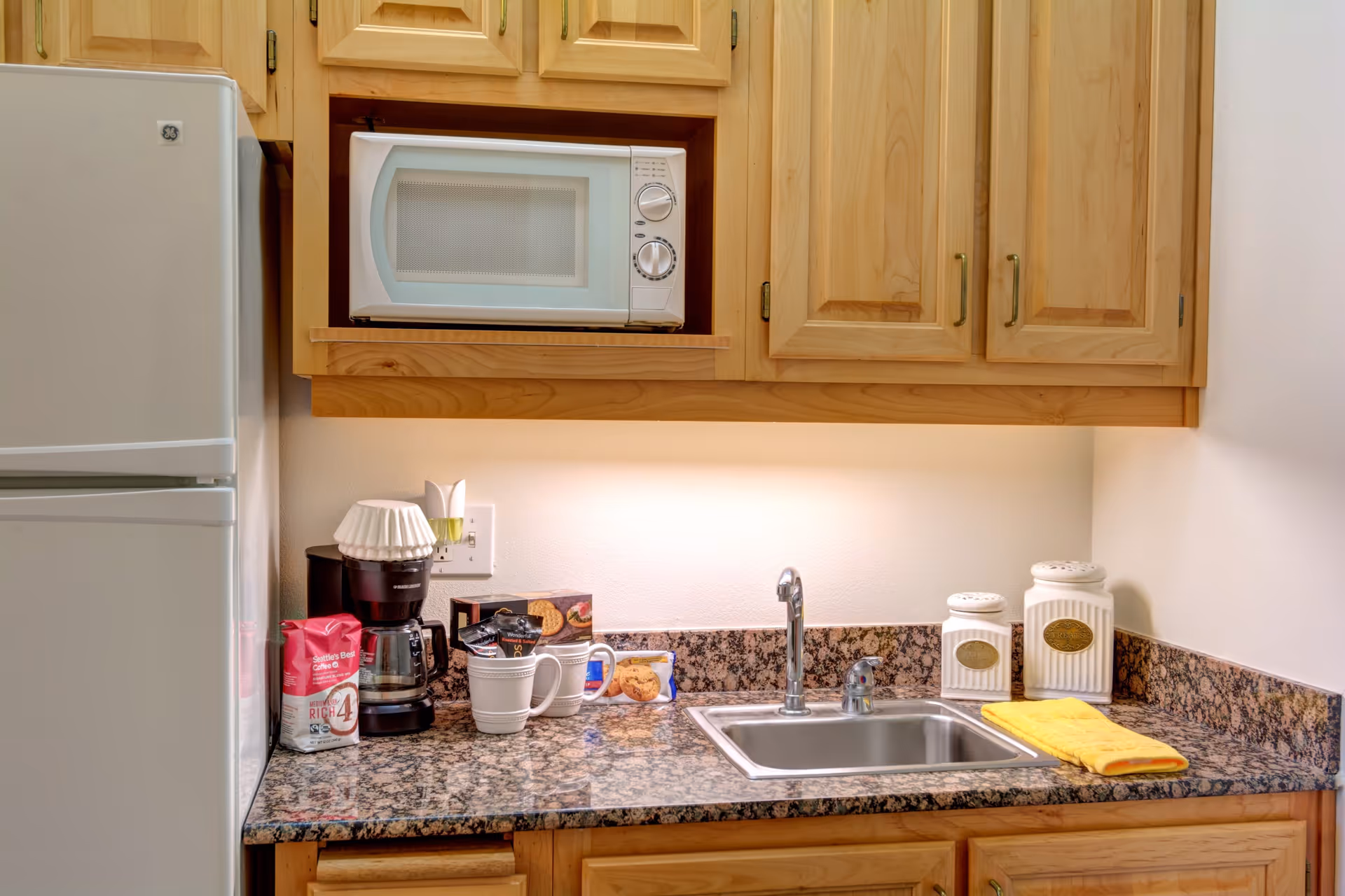 A small kitchen area with wooden cabinets, a white microwave, a coffee maker with coffee mugs, a granite countertop with a sink, and a refrigerator on the left side. There are also two white canisters and a yellow towel on the countertop.