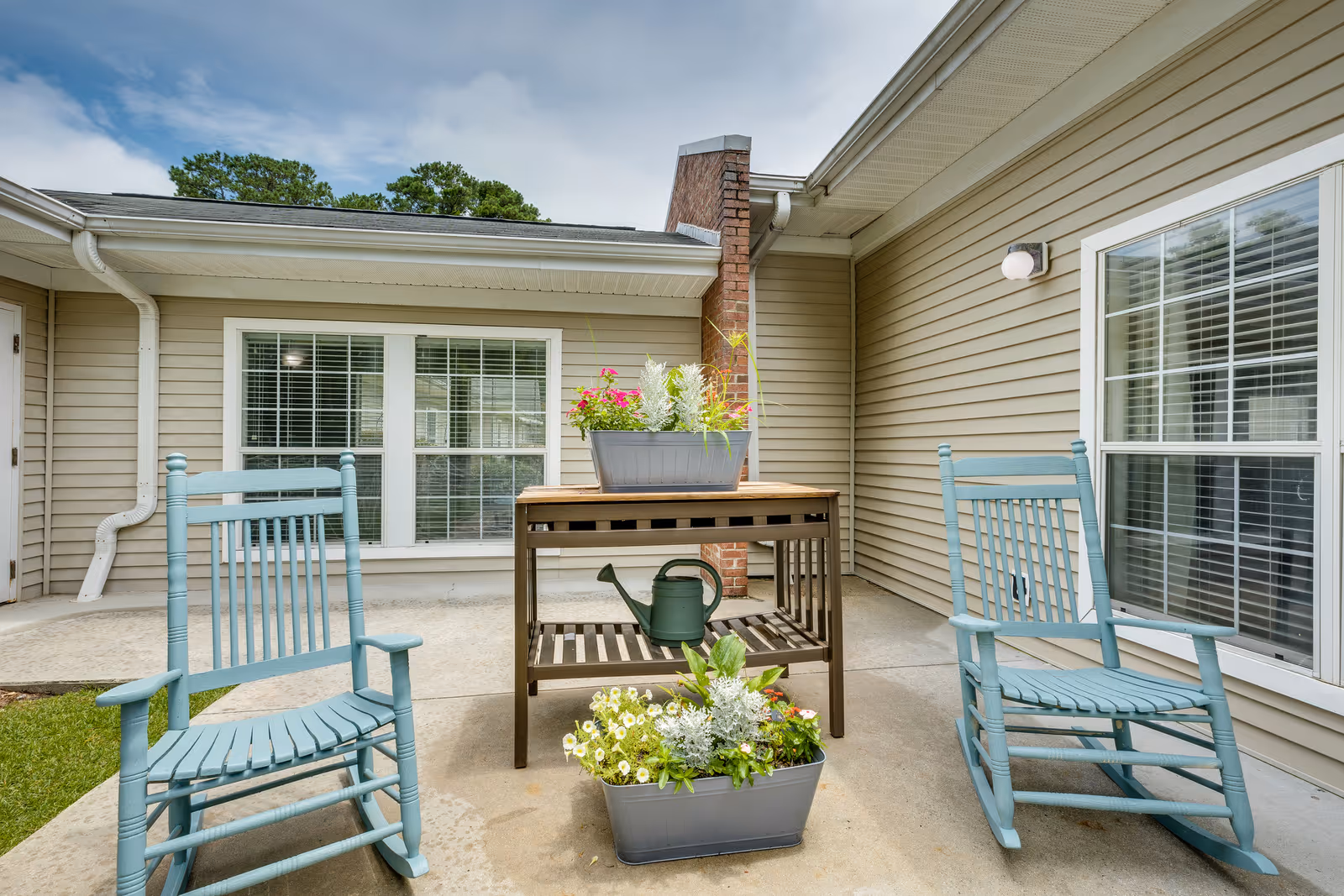 Outdoor patio area with two light blue wooden rocking chairs facing each other, a small wooden table with a watering can on the lower shelf, and two rectangular planters filled with flowers and greenery. The patio is adjacent to a beige siding building with white-framed windows and a brick chimney.
