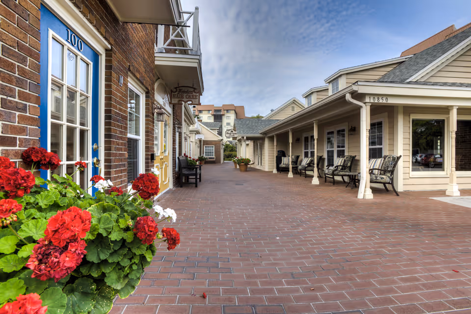 Outdoor walkway area of a senior living facility with brick flooring, red and white flowers in planters, benches along the walls, and buildings with windows and doors on either side under a partly cloudy sky.