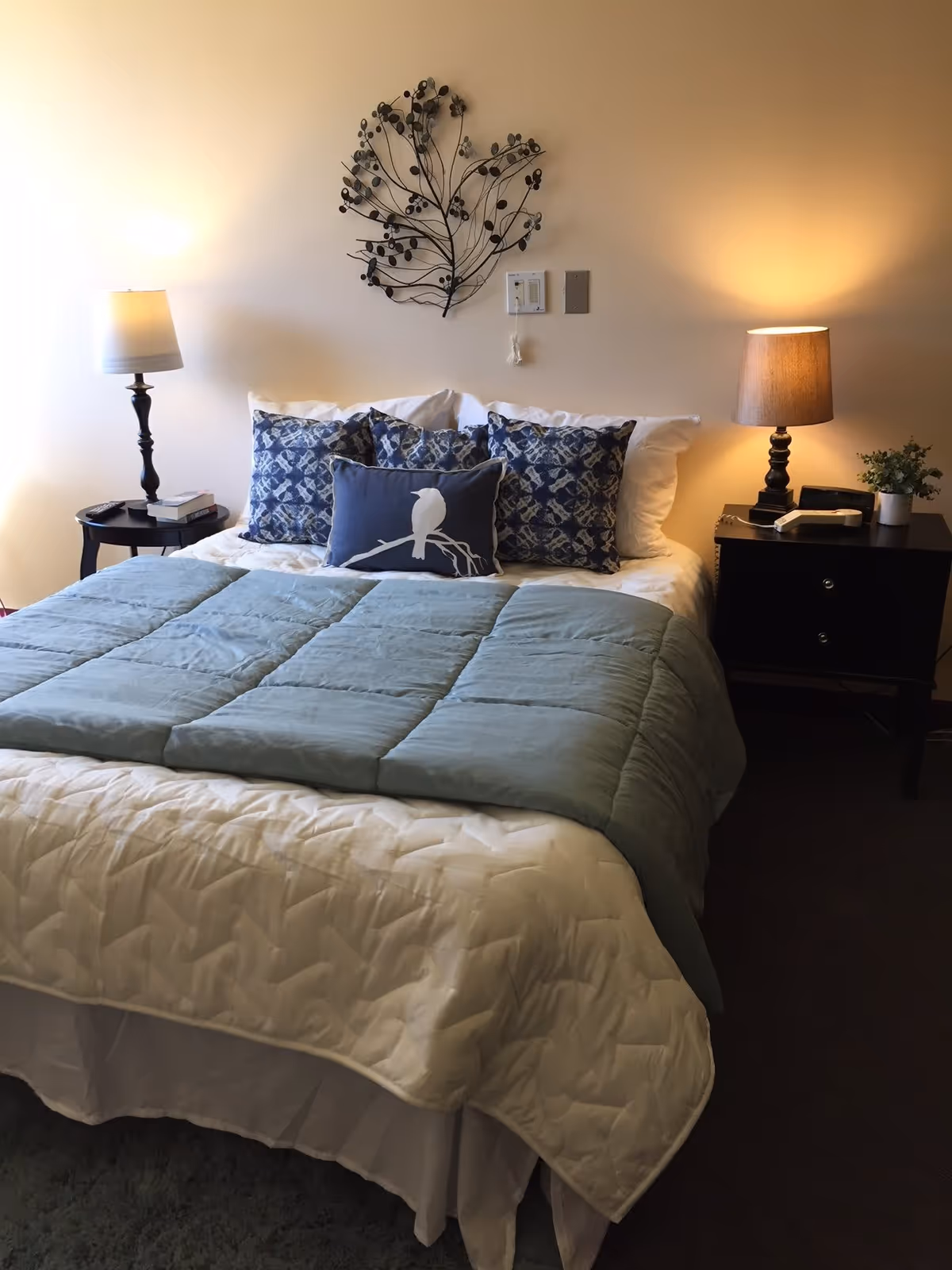 A neatly made bed with a white quilt and a light blue comforter folded at the foot. The bed has four decorative pillows, three with a blue and white pattern and one with a bird design. On either side of the bed are dark wooden nightstands with lamps; the left lamp has a white shade and the right lamp has a beige shade. Above the bed is a decorative metal wall art piece shaped like branches with berries. There is a small plant and a telephone on the right nightstand.