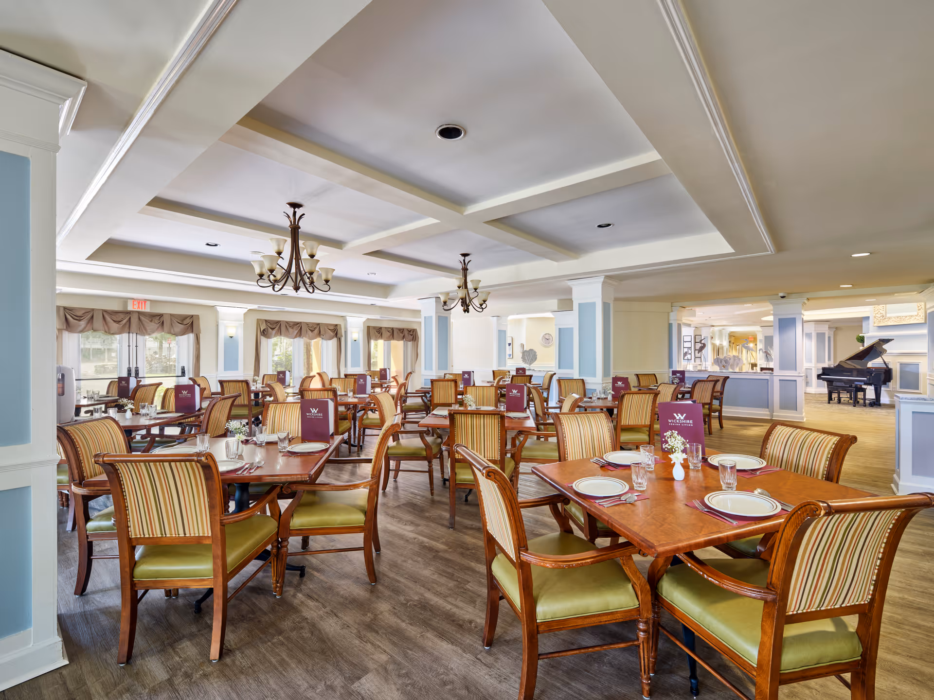 A spacious dining room with multiple wooden tables and striped cushioned chairs arranged neatly. Each table is set with plates, glasses, silverware, and a small flower vase. Menus labeled 'Wickshire Tamarac' are placed on the tables. The room features coffered ceilings with chandeliers, large windows with valances, and a grand piano in the background.