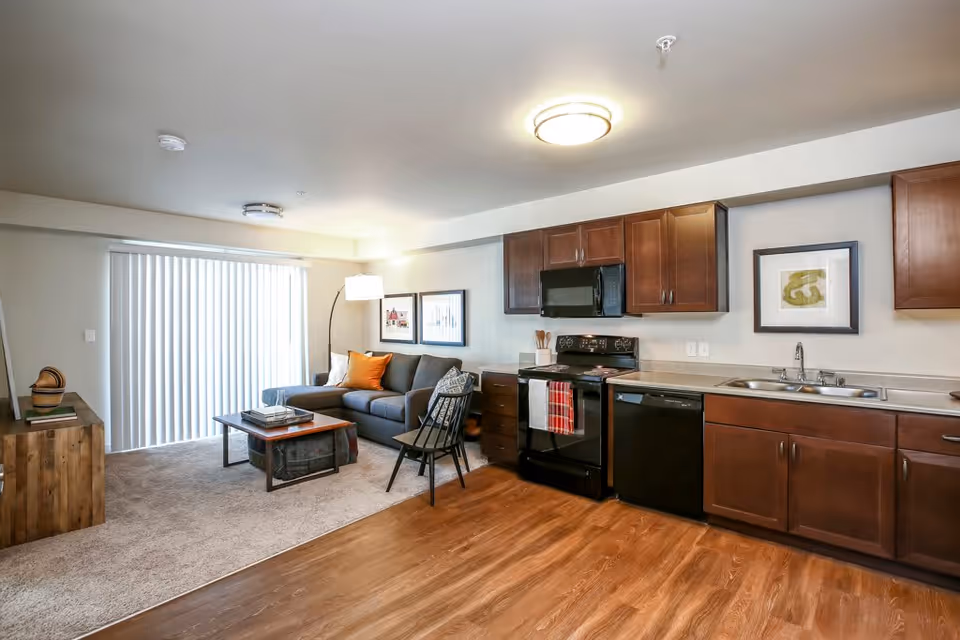 Open-concept living area and kitchen in a senior living facility apartment. The living area features a gray sofa with orange and patterned pillows, a wooden coffee table, a black chair, and a floor lamp. The kitchen has dark wood cabinets, a black stove, microwave, dishwasher, and a double sink. The floor transitions from carpet in the living area to wood in the kitchen. Sliding vertical blinds cover a large window or door in the background.