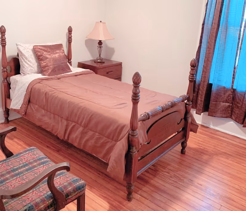 A small bedroom with a wooden single bed covered with a brown comforter and pillows. Next to the bed is a wooden nightstand with a lamp. There is a wooden chair with a patterned cushion in the foreground and a window with dark curtains letting in some light.