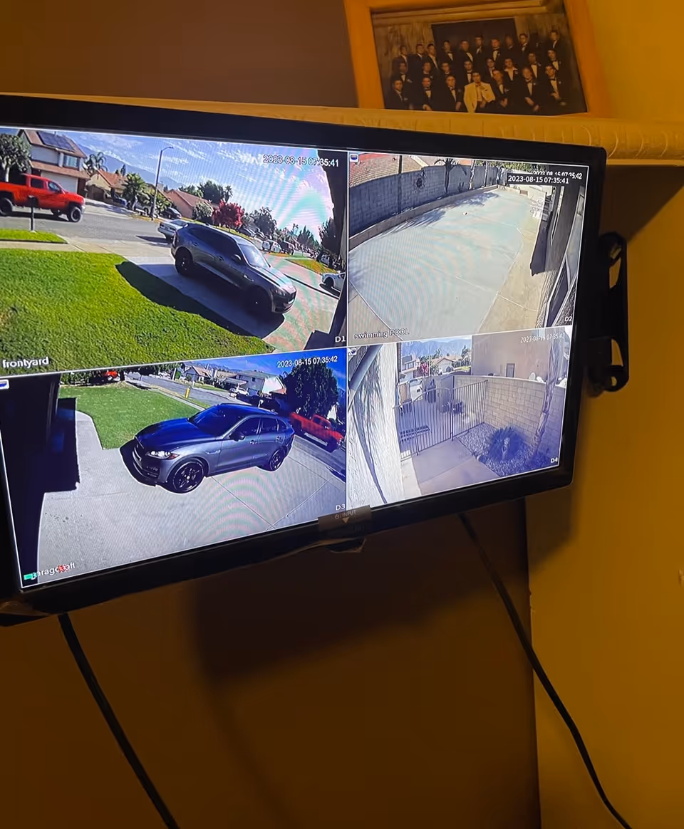 A wall-mounted monitor displaying four security camera views showing different outdoor areas including a front yard with a parked black SUV, a driveway with another black vehicle, a concrete pathway, and a gated patio area. The monitor is mounted on a yellow wall with a framed group photo above it.