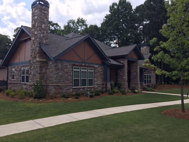 Front exterior of a single-story stone-clad building with a chimney, lawn, and a sidewalk.