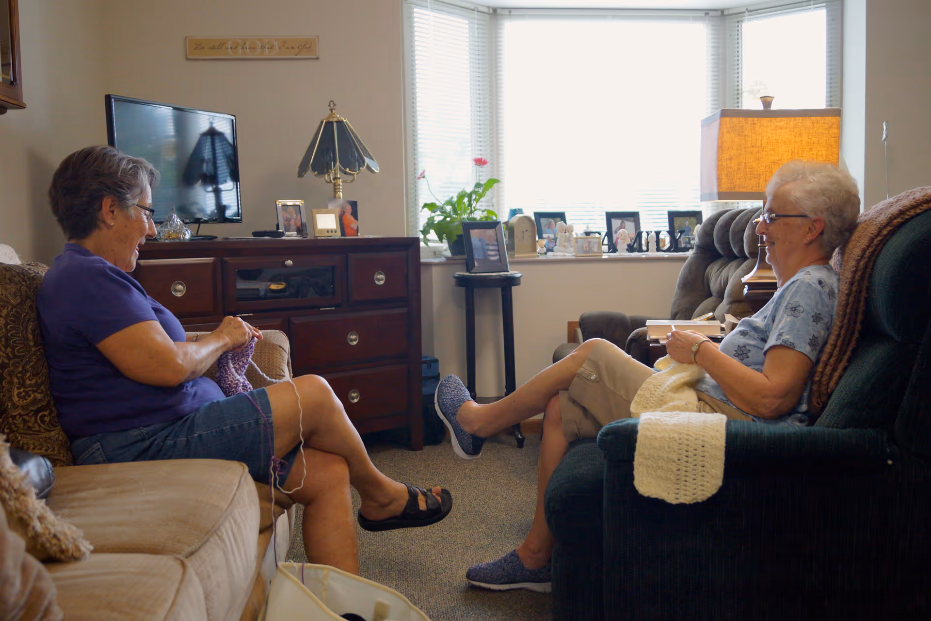 Two elderly women sitting in a cozy living room, facing each other and knitting. One woman is seated on a beige couch wearing a purple shirt and denim shorts, while the other is on a green armchair wearing a light blue shirt and beige pants. The room has a wooden cabinet with a TV, a table lamp, framed photos, and a window with blinds letting in natural light.
