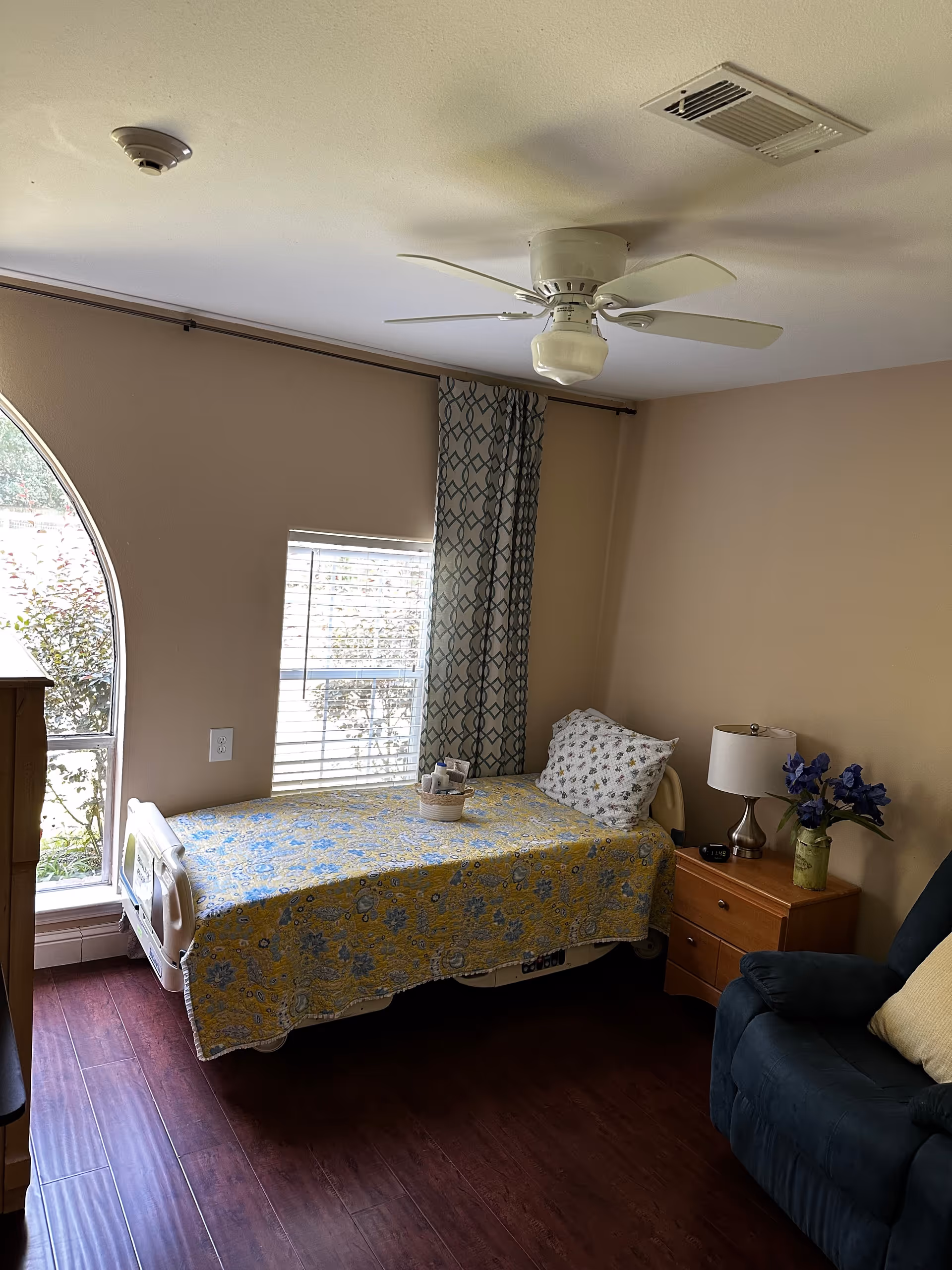 A cozy bedroom in a senior living facility with a single bed covered in a yellow and blue floral quilt. The bed is positioned near a window with patterned curtains and an arched window beside it. Next to the bed is a wooden nightstand with a lamp, a vase with purple flowers, and a small clock. A dark blue armchair with a yellow pillow is partially visible on the right side. The room has wooden flooring and a ceiling fan with a light fixture.