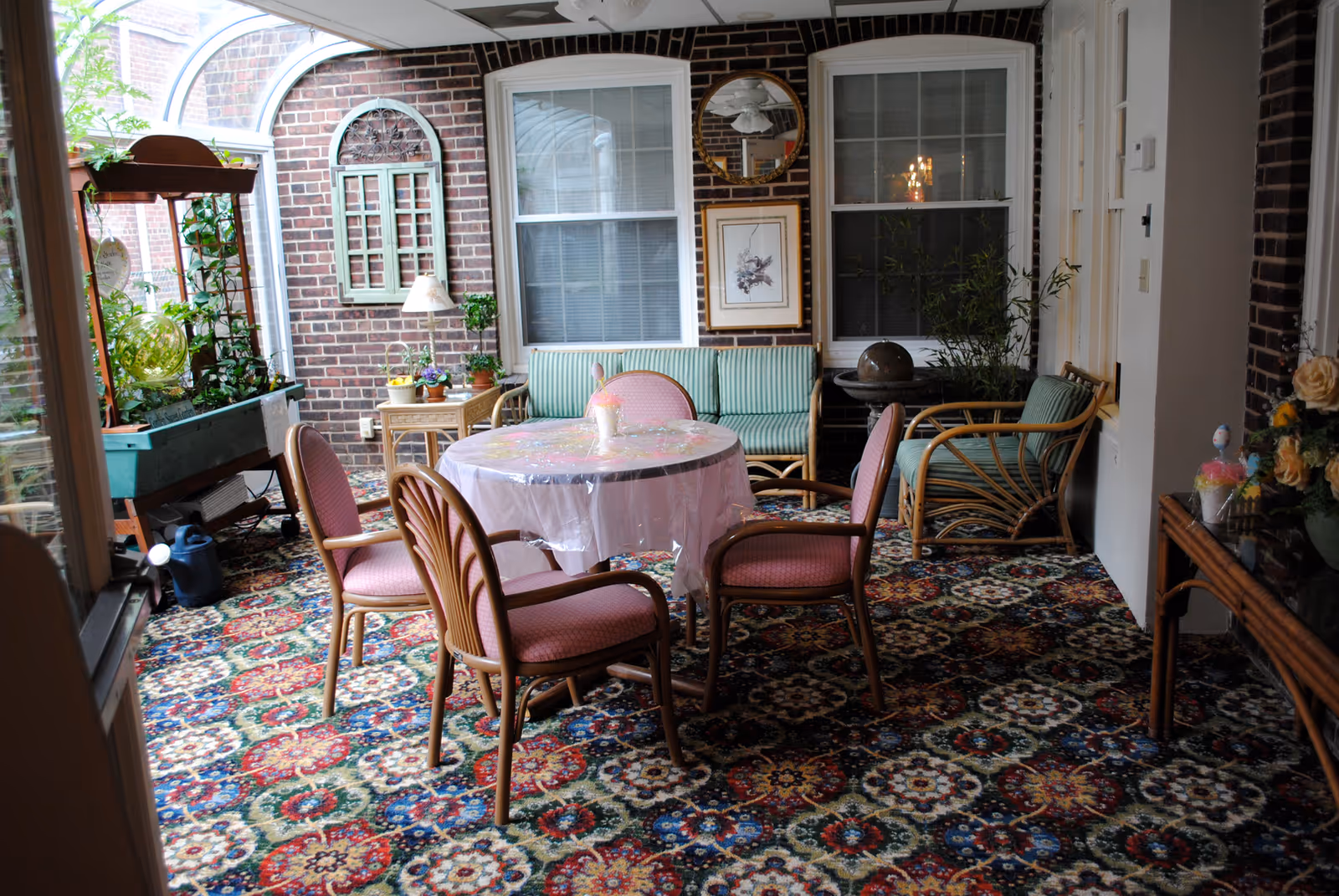A cozy indoor sitting area with a round table covered with a plastic tablecloth surrounded by four pink cushioned chairs. Behind the table, there is a green striped cushioned sofa and a matching armchair. The room has a colorful patterned carpet and brick walls with two large windows. There are various plants and decorative items, including a round mirror and framed artwork on the wall.