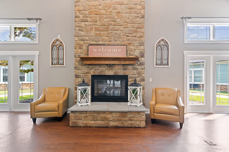 A cozy living room area featuring a stone fireplace with a wooden mantel. On the mantel is a sign that says 'welcome'. Two beige armchairs are placed symmetrically on either side of the fireplace. Each side of the fireplace has a decorative lantern on the hearth and a wall decoration above the chairs. Large windows and glass doors on both sides allow natural light to fill the room, showing a view of the outside greenery.
