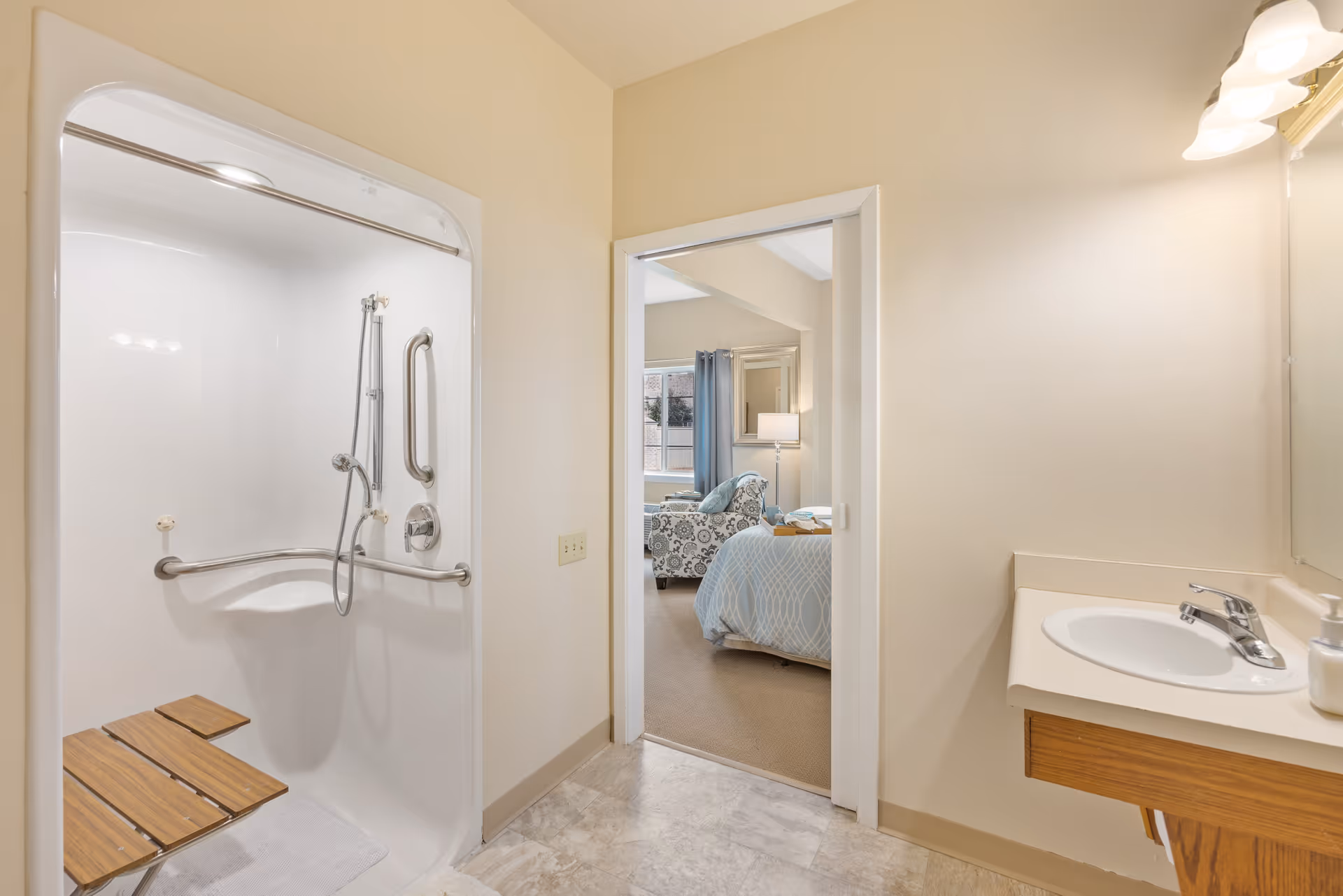 A bathroom with a walk-in shower featuring a fold-down wooden seat and grab bars. The bathroom has a sink with a faucet and soap dispenser on the right. Through an open doorway, a bedroom is visible with a bed covered in a light blue patterned blanket, a patterned armchair, a floor lamp, and a window with curtains.