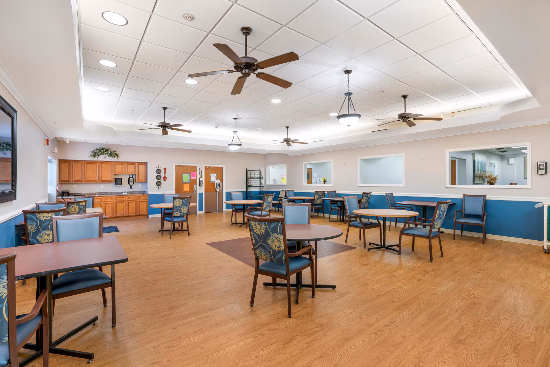A spacious dining room with multiple round and rectangular tables surrounded by blue cushioned chairs with floral patterns. The room has a wooden floor, ceiling fans, and hanging light fixtures. There is a kitchenette area with wooden cabinets and a countertop along one wall, and several windows and doors are visible.