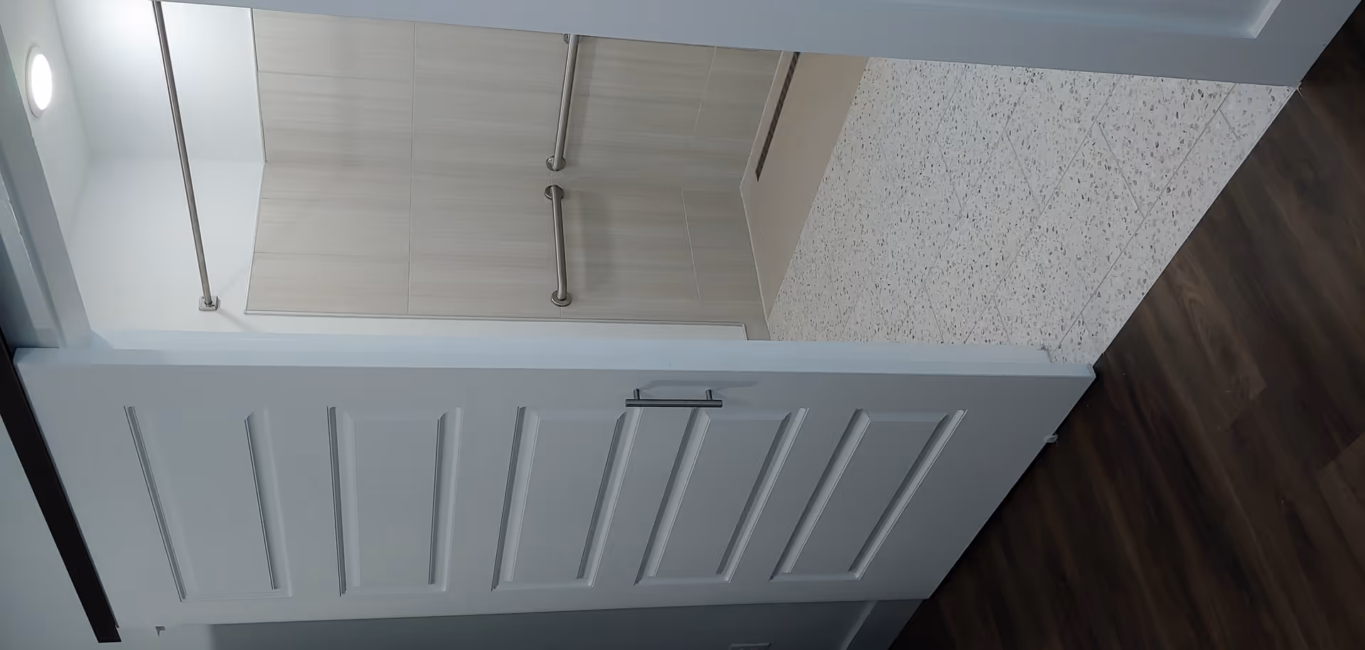 View of a bathroom entrance with a white sliding door partially open, revealing a tiled shower area with two horizontal grab bars and a shower curtain rod. The floor outside the bathroom is dark wood, and the bathroom floor is light-colored tile.