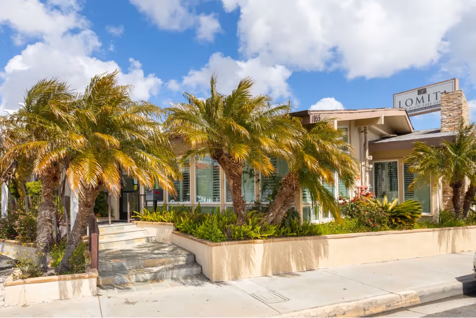 Exterior view of Lomita Post-Acute Care Center building with palm trees and greenery in front, a stone staircase leading to the entrance, and a partly cloudy blue sky above.